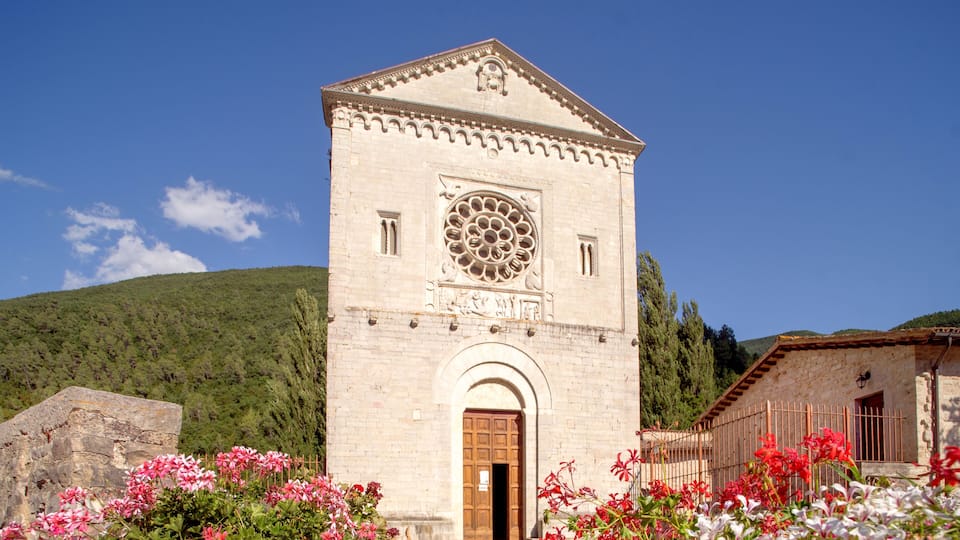 A historic church with a large rose window, surrounded by blooming flowers and set against a mountainous backdrop.