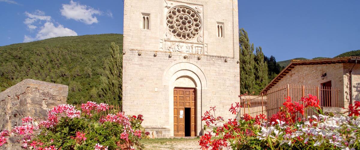 A historic church with a large rose window, surrounded by blooming flowers and set against a mountainous backdrop.