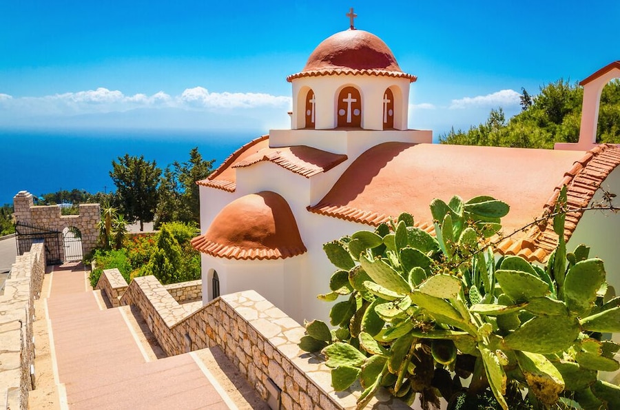 Typical Greek church with red roof and green plants in front and sea view in the background. Greece; Shutterstock ID 313454183; PO: Hcom Destination Content neighborhoods; Client: Hotels.com; Other: H