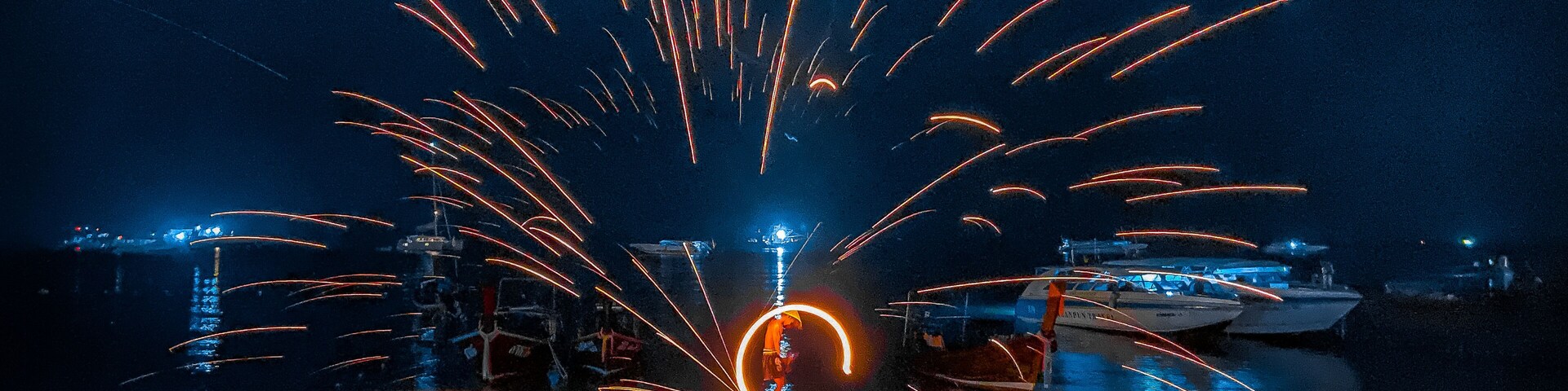 Cena noturna de praia com pessoas sentadas ao redor de uma fogueira, barcos ao fundo e um espetáculo de fogos de artifício.