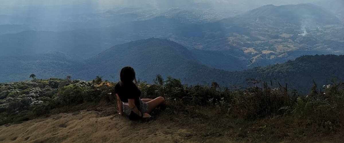 A person sitting on a hill overlooking a valley with mountains in the distance and clouds in the sky.