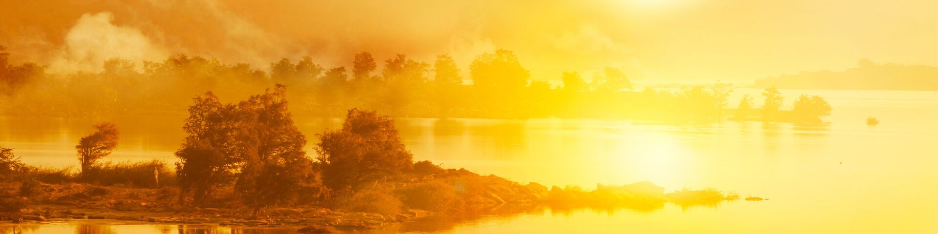 Nascer do sol sobre um lago calmo, com árvores e montanhas ao fundo.