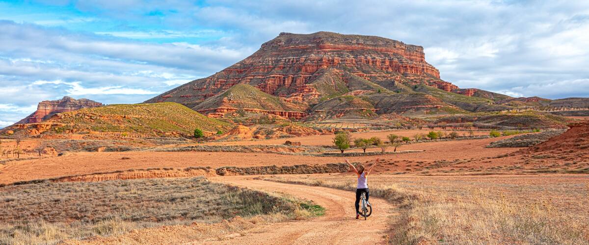 A person riding a bicycle on a dirt road with a large red rock formation in the background.