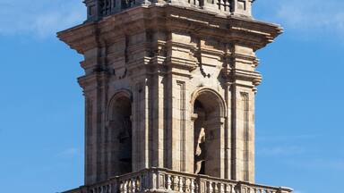 A tall stone tower with a dome and a cross on top, featuring intricate carvings and a bell.