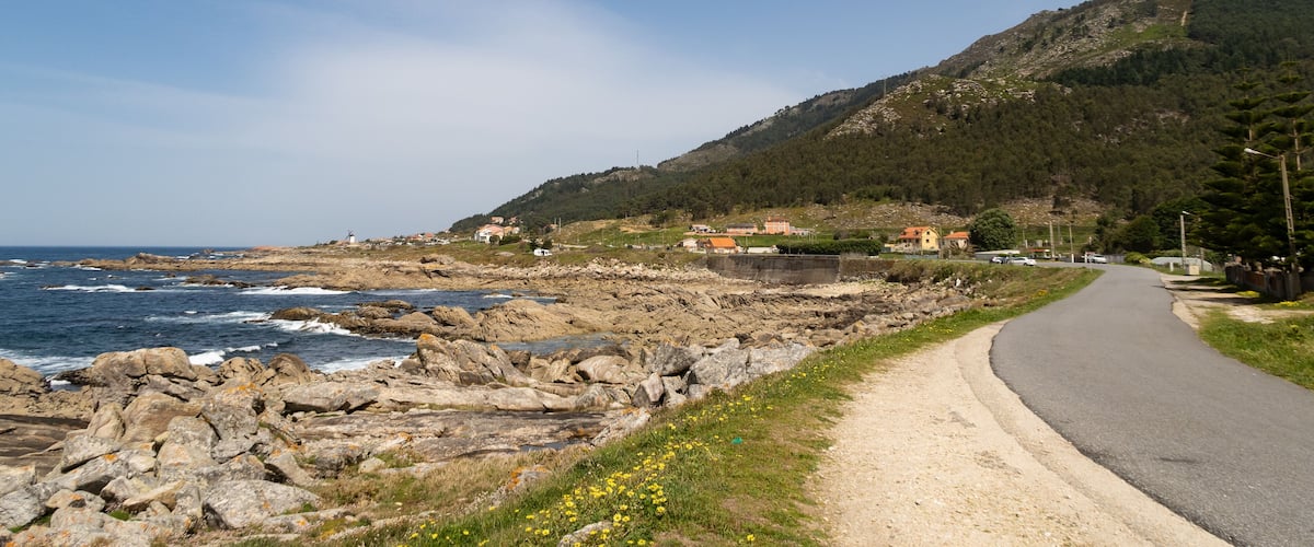 A coastal road with rocky terrain, a clear sky, and a hill with buildings in the background.