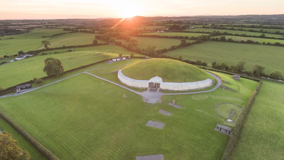 A circular mound with a central structure, surrounded by a field and a few buildings.