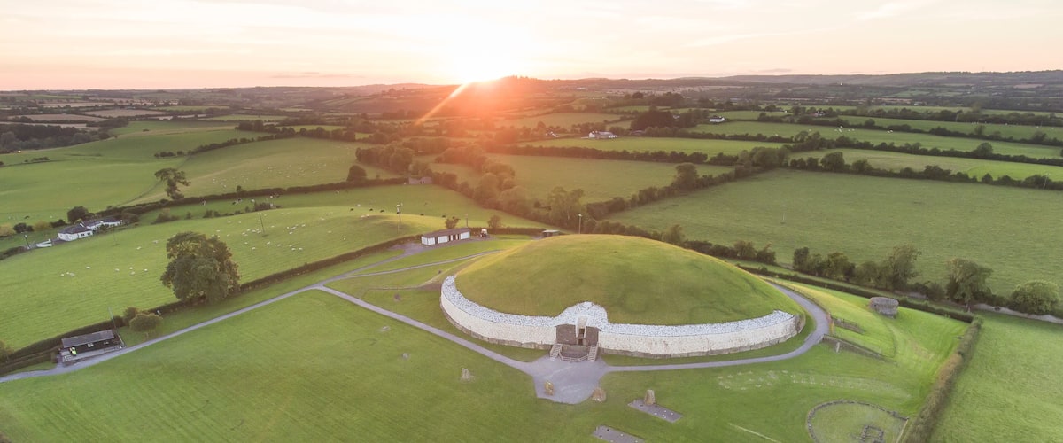 A circular mound with a central structure, surrounded by a field and a few buildings.