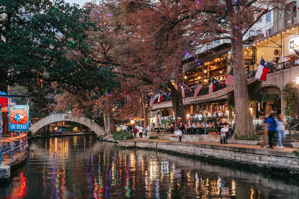 Um rio com uma ponte de pedra, árvores com luzes coloridas e um restaurante com bandeiras.