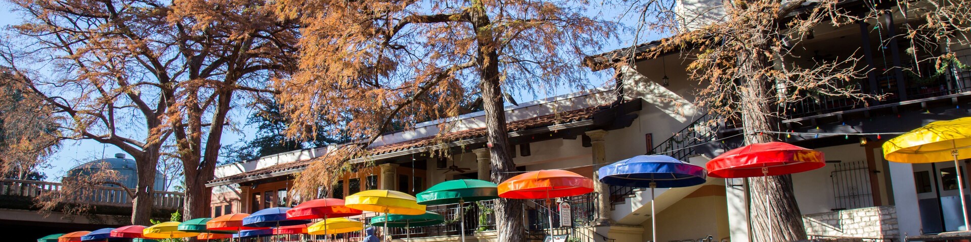 Un café a la orilla del río con sombrillas de colores y árboles otoñales.