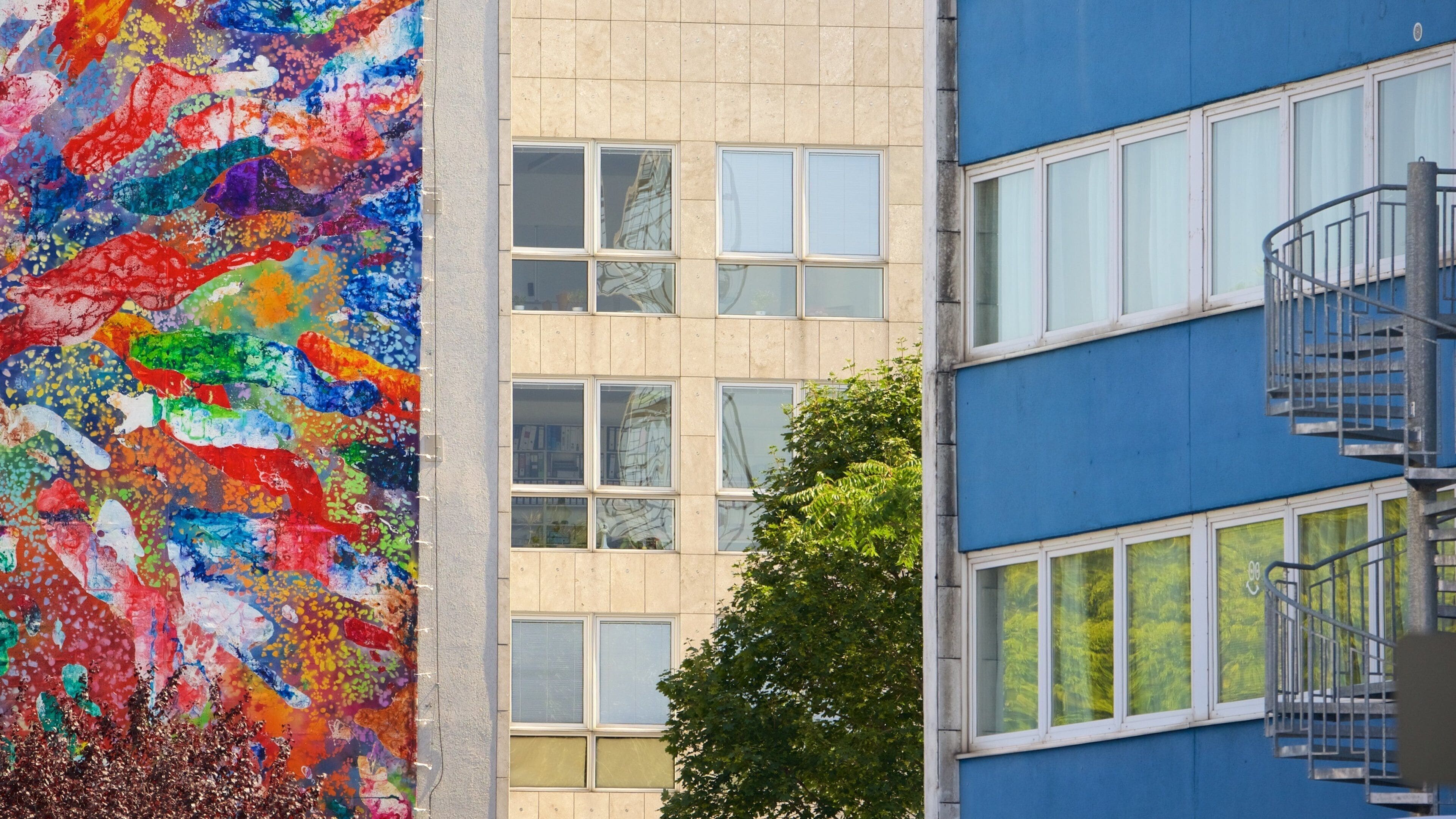 Une fresque colorée orne un bâtiment, qui est une structure beige dotée de nombreuses fenêtres, et un autre bâtiment, de couleur bleue, est équipé d’un escalier en métal.