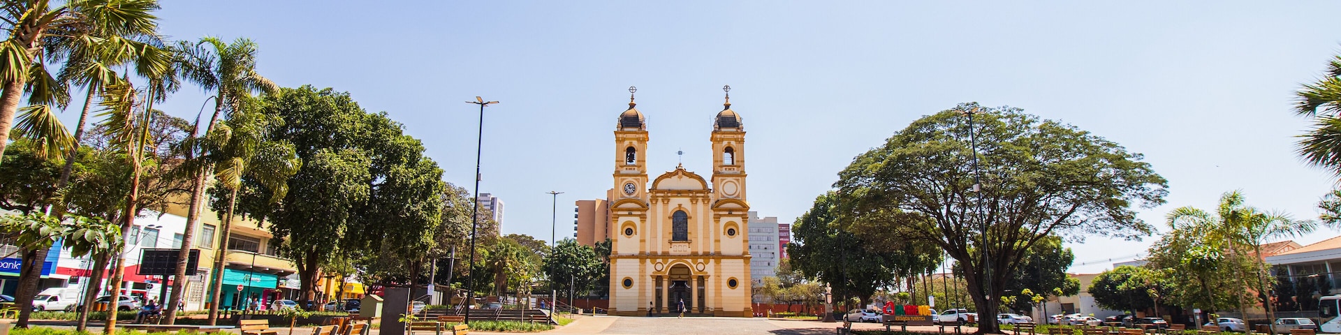 A plaza with a historic church, palm trees, benches, and a clear blue sky.