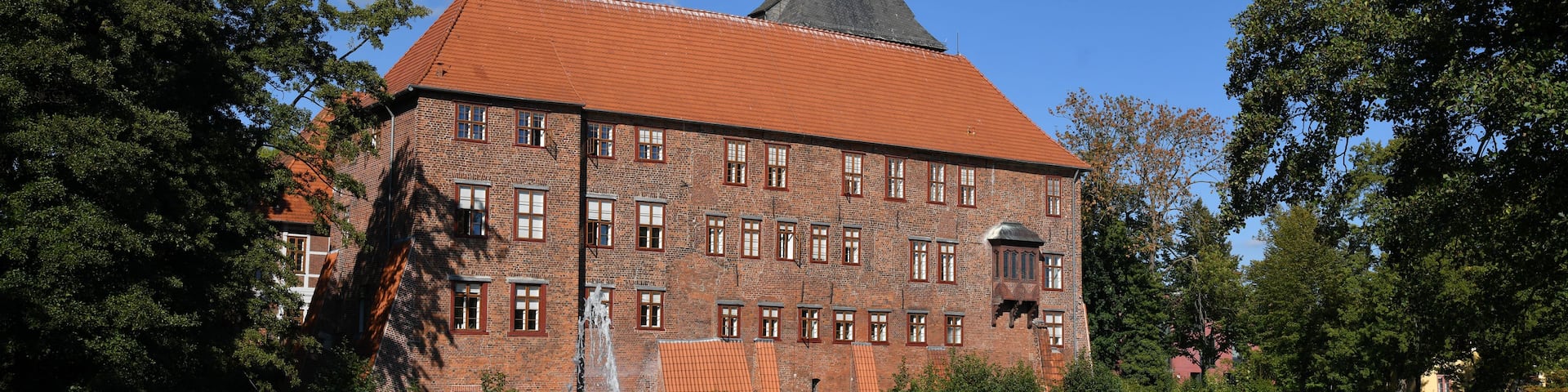 A historic building with a red-tiled roof and a tower, surrounded by trees and a body of water with a fountain.