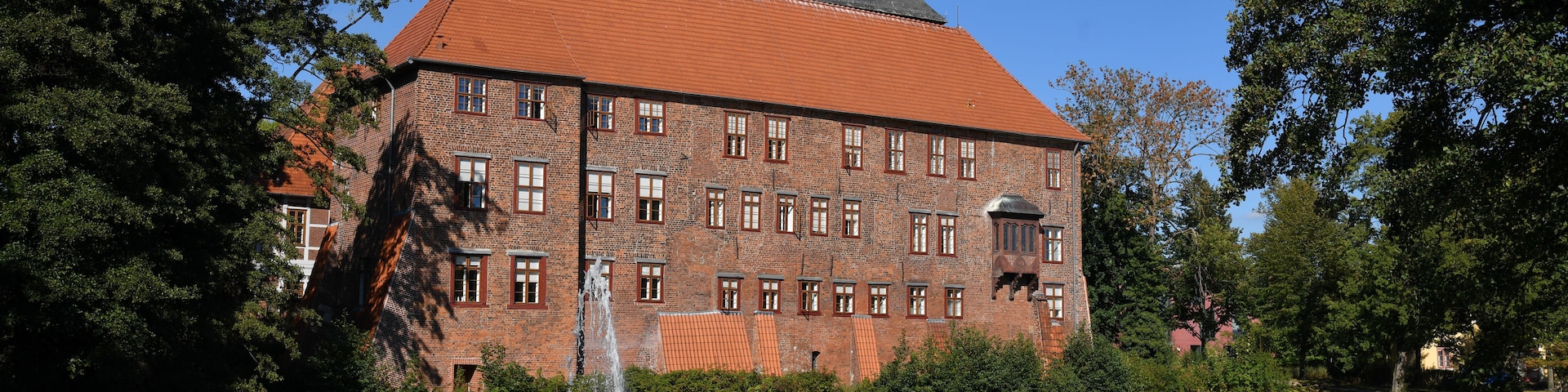 Un edificio histórico con techo de tejas rojas y una torre, rodeado de árboles y un cuerpo de agua con una fuente.