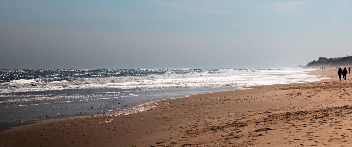A sandy beach with waves crashing onto the shore and a few people walking along the beach.