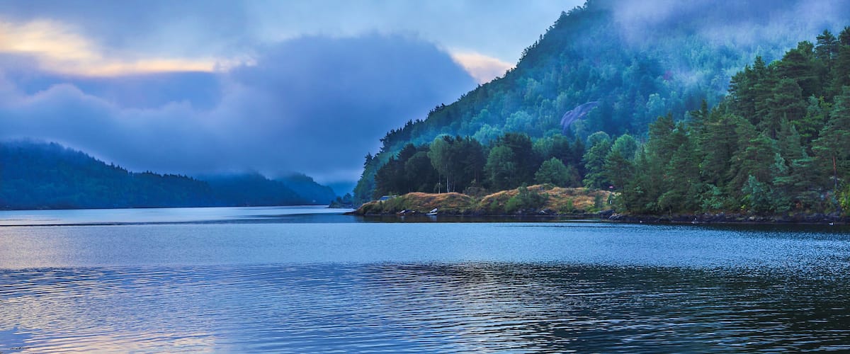 A calm lake surrounded by dense forests and mist-covered mountains.