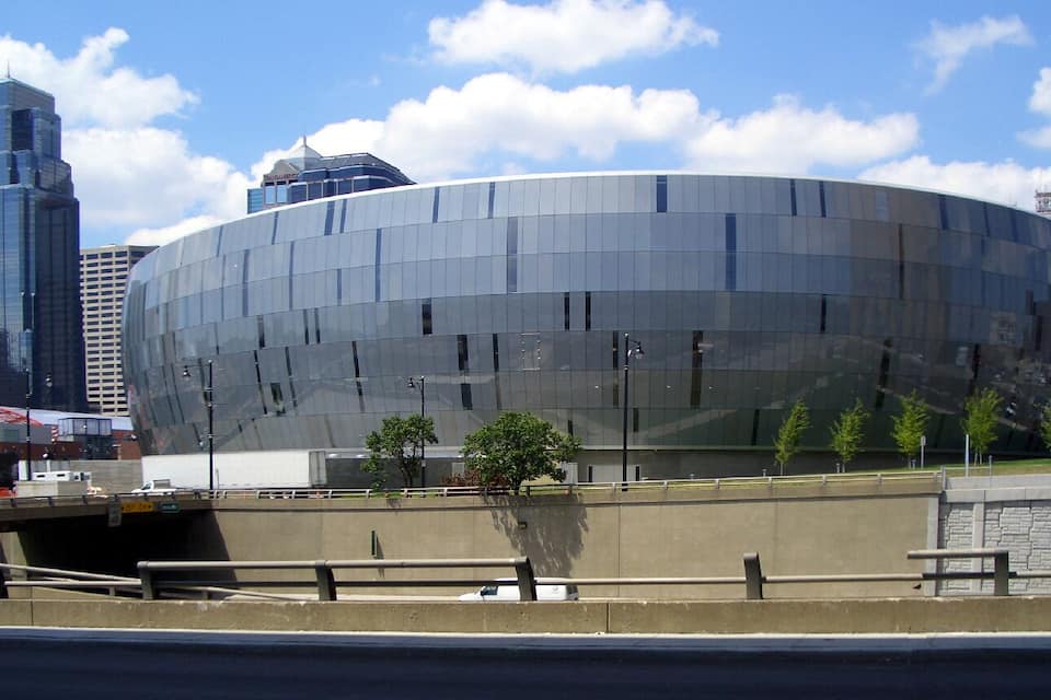 Een modern, gebogen glazen gebouw met een reflecterende gevel, omgeven door een betonnen barrière en enkele bomen.