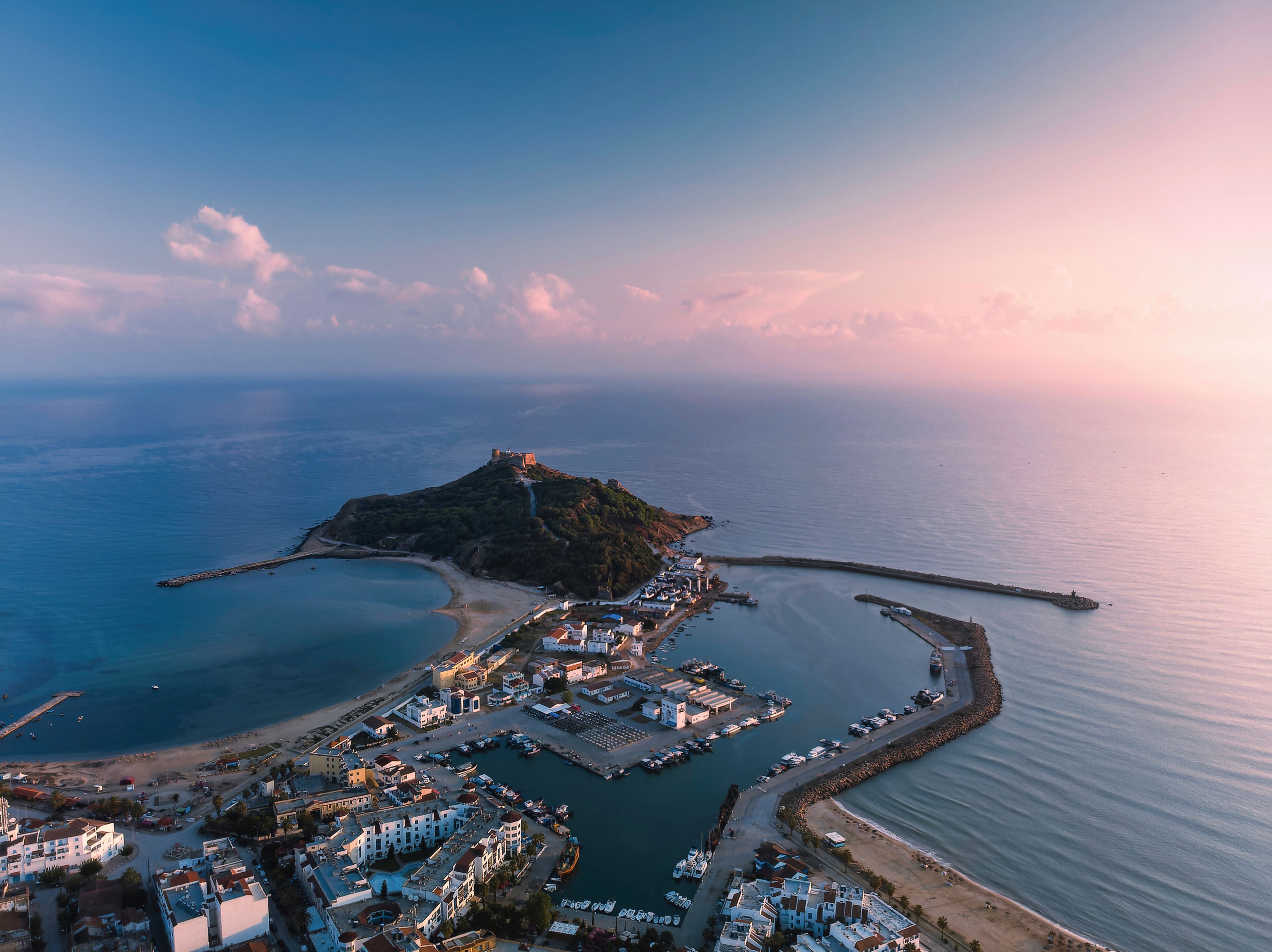 An aerial view of a coastal town with a prominent hilltop castle, a marina, and a beach.