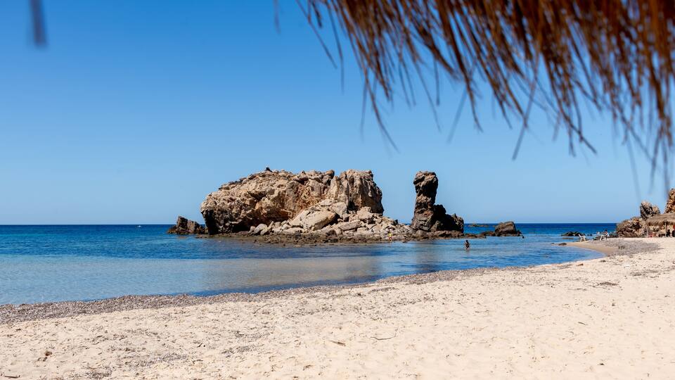 A sandy beach with clear blue water, rocky formations, and a clear sky.