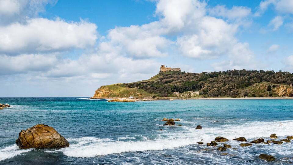 A coastal landscape with a castle on a hill, clear blue sky, and rocky shoreline.