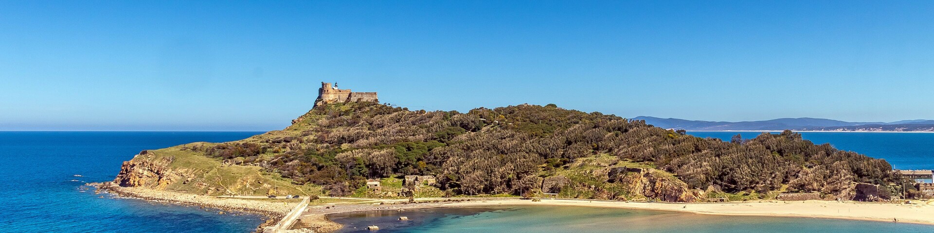 Un paisaje costero con una colina coronada por un castillo, aguas azul cristalino y una playa de arena.
