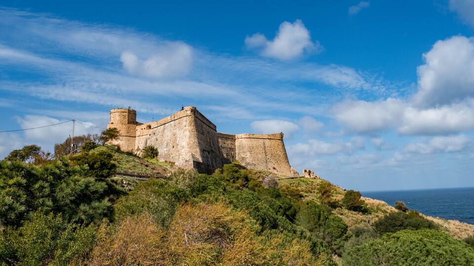 A historic stone fortress on a hill with a clear sky and the sea in the background.