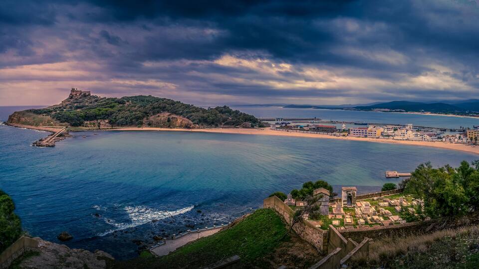 A coastal landscape with a beach, a fortified hilltop, and a small cemetery.