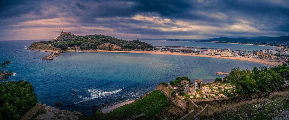 A coastal landscape with a beach, a fortified hilltop, and a small cemetery.
