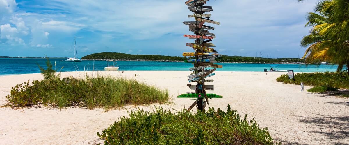 A beach with a tall signpost with multiple directional signs, a clear blue sky, and a boat in the water.