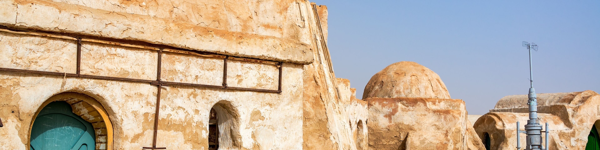 Paisagem desértica com estruturas de pedra antigas, incluindo um edifício com porta azul e janelas em arco, e um grande monte ao fundo.