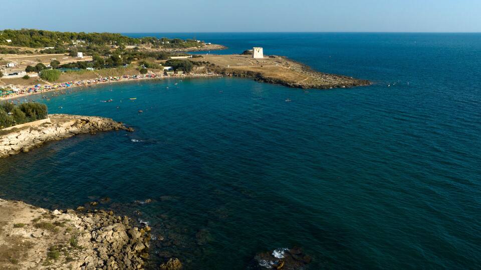 A coastal landscape with a rocky shoreline, a beach with umbrellas, and a tower on a small island.