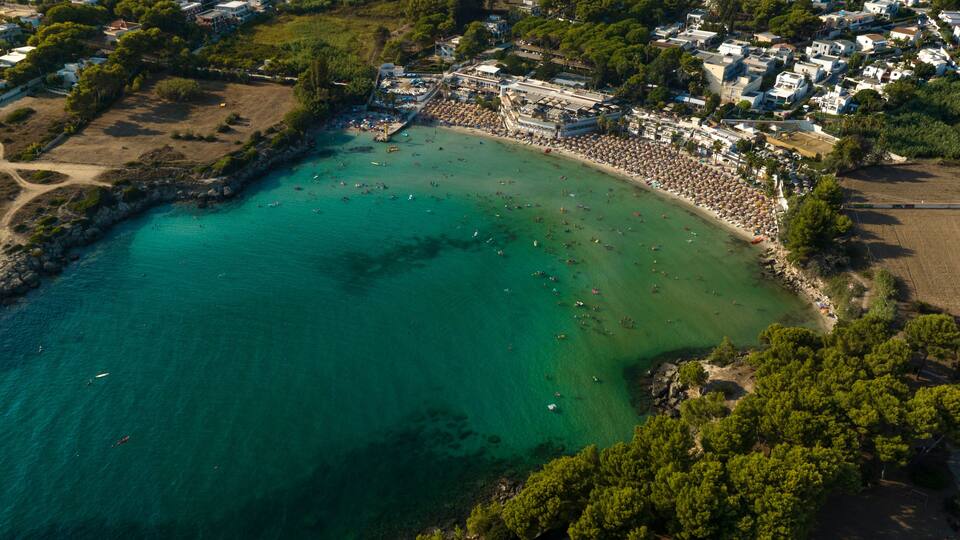 An aerial view of a coastal area with a beach, clear turquoise waters, and surrounding buildings.