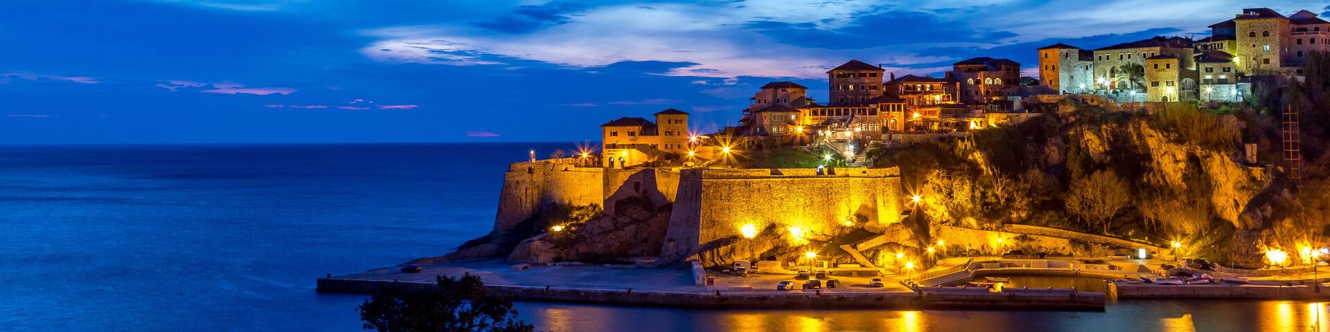 A coastal town at dusk with illuminated buildings and a fortified structure overlooking the sea.