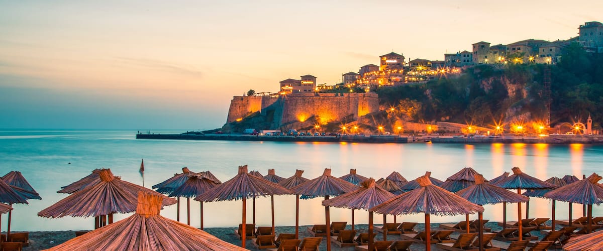 A beach with rows of wooden lounge chairs and thatched umbrellas, a calm sea, and a historic fortified hill with buildings in the background.
