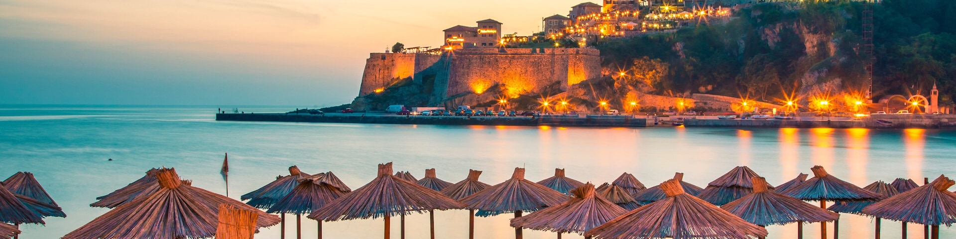 A beach with rows of wooden lounge chairs and thatched umbrellas, a calm sea, and a historic fortified hill with buildings in the background.