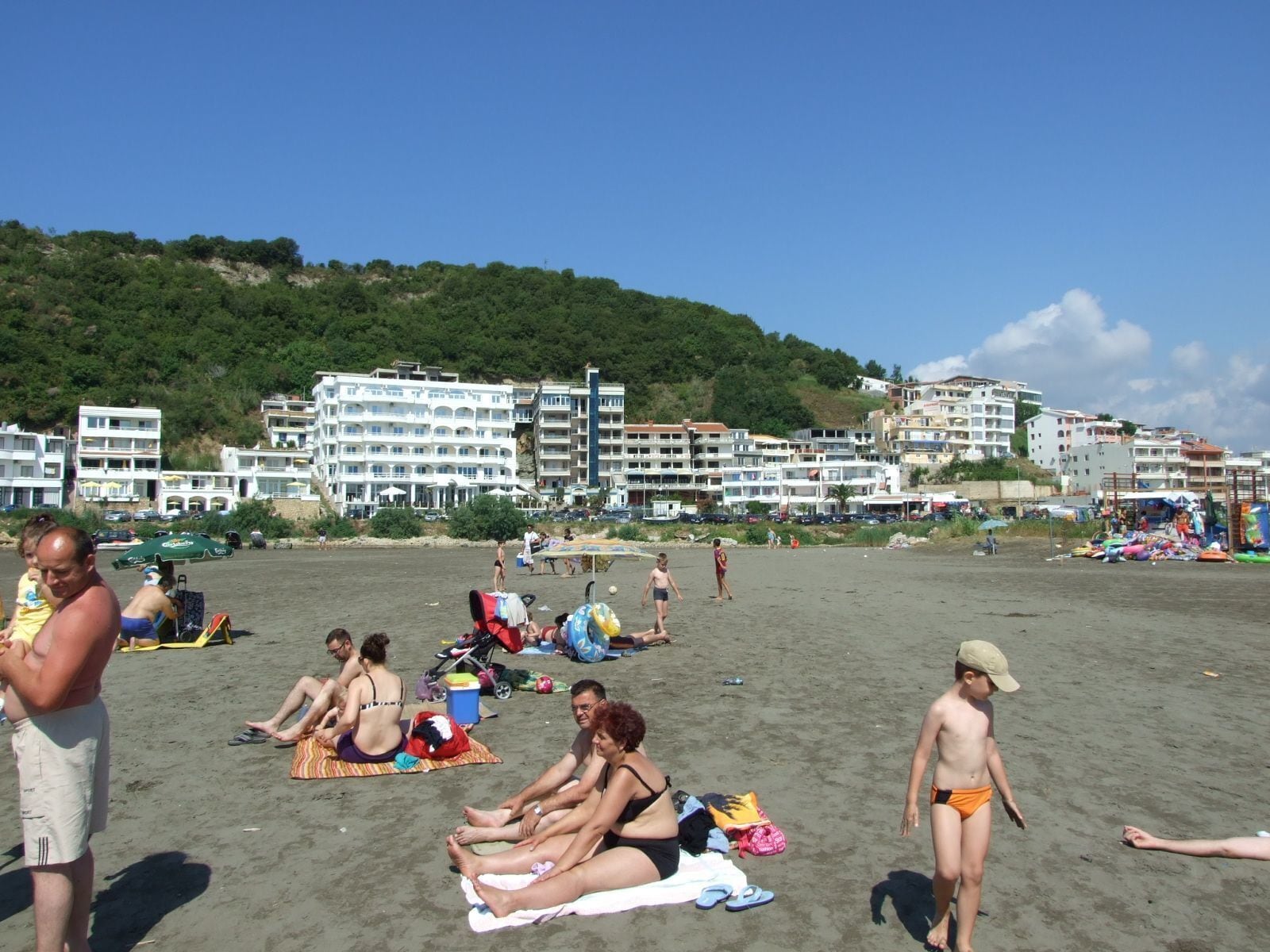 A beach with people sunbathing and walking, surrounded by buildings and hills.