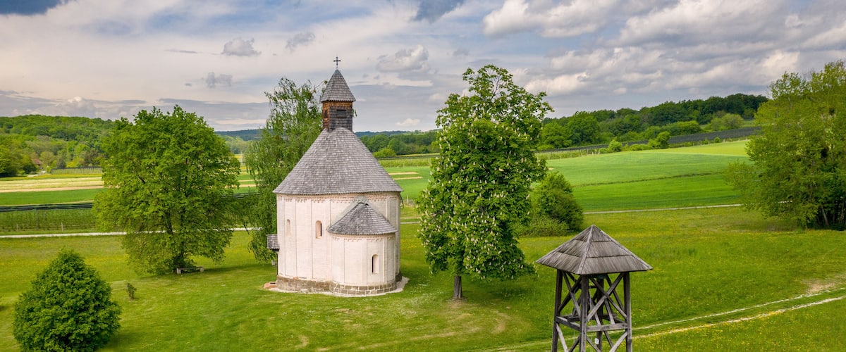 A white church with a bell tower and a wooden gazebo in a lush green field.