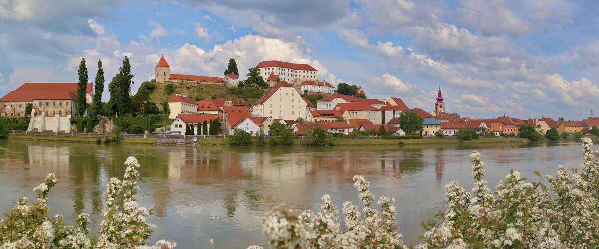 A historic town with a castle on a hill, a river in the foreground, and traditional buildings with red roofs.