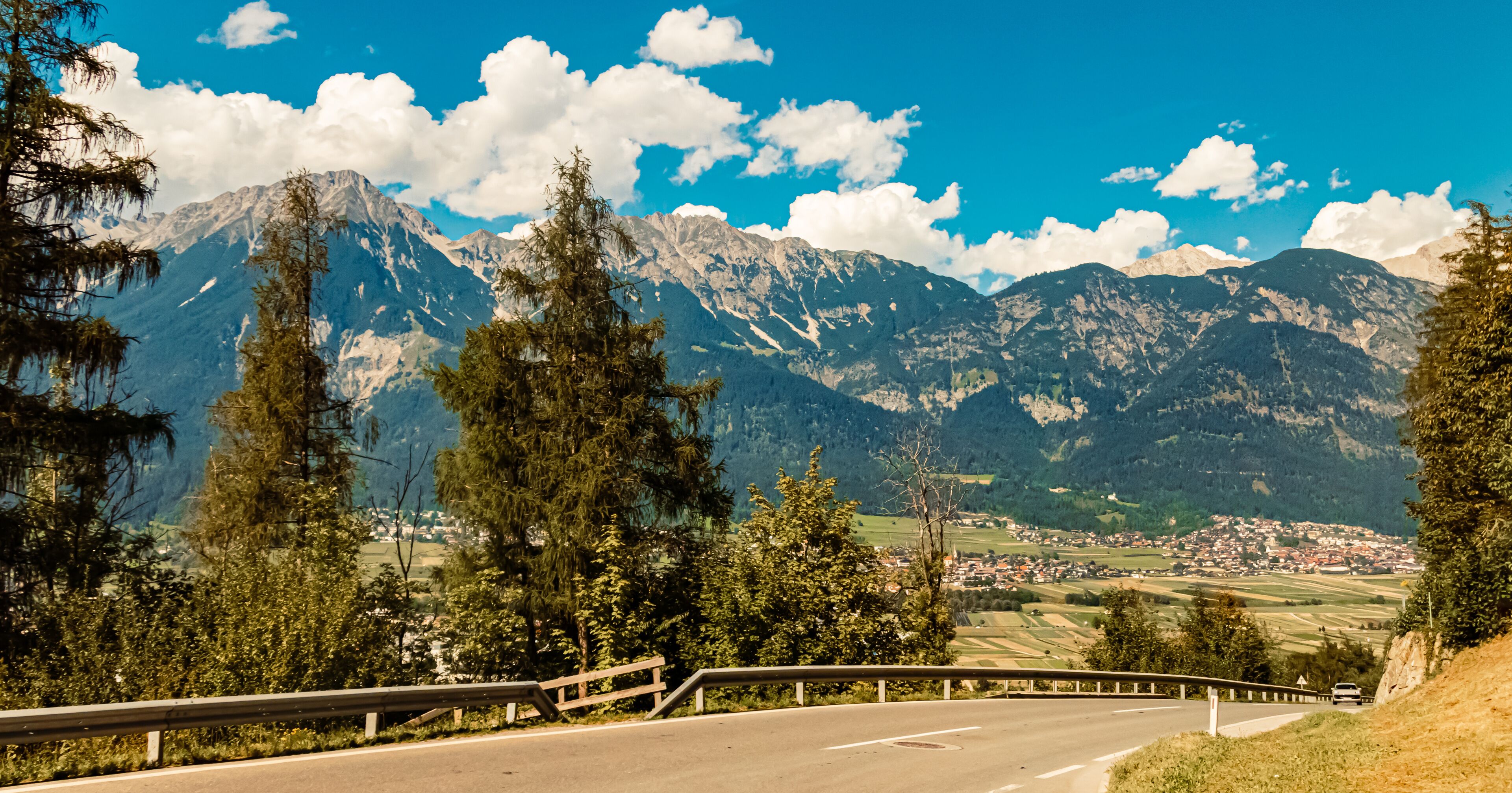 A winding road with a guardrail, surrounded by tall evergreen trees and a mountain range with a small town in the valley below.
