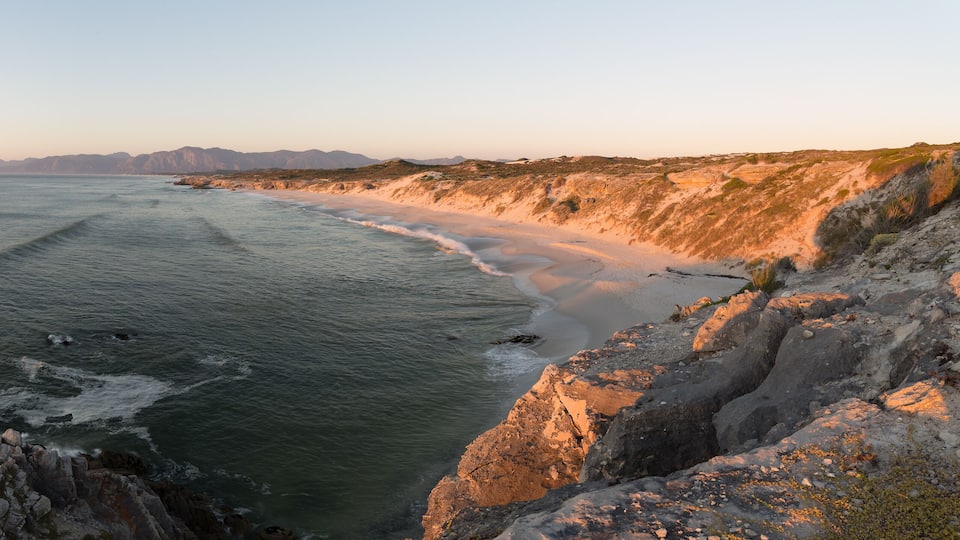 A coastal landscape with a rocky shoreline, waves crashing against the rocks, and distant mountains under a clear sky.