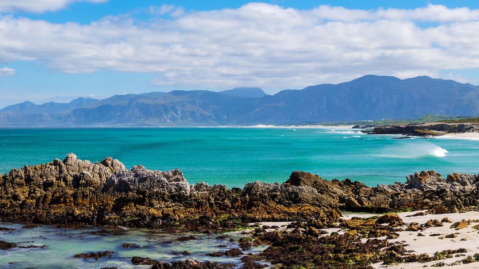 A coastal landscape with rocky formations, clear turquoise waters, and distant mountains.