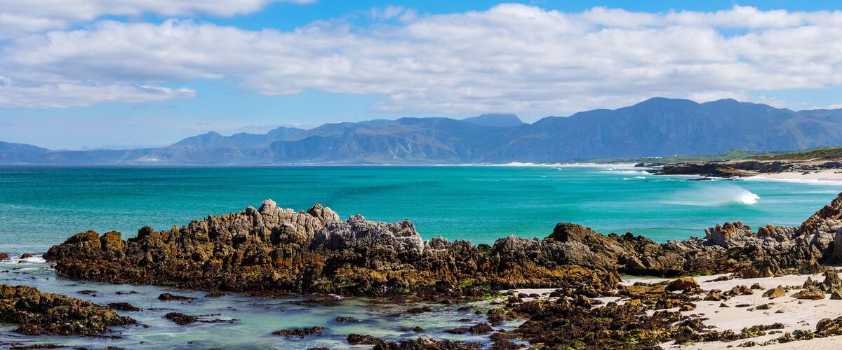 A coastal landscape with rocky formations, clear turquoise waters, and distant mountains.