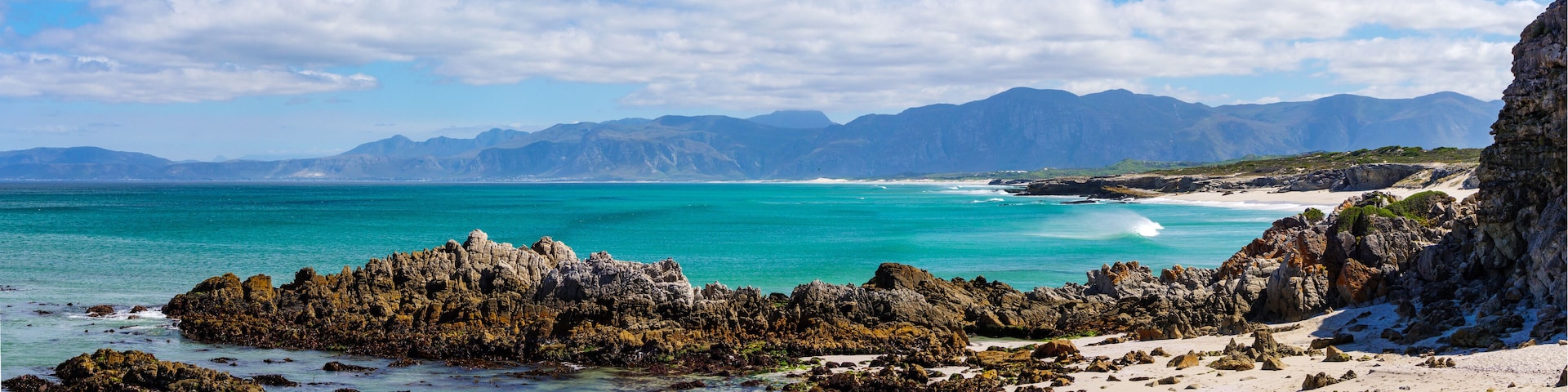 A coastal landscape with rocky formations, clear turquoise waters, and distant mountains.