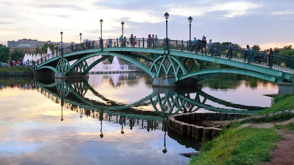 A green bridge over a calm body of water with reflections of the sky and bridge, surrounded by greenery and a distant cityscape.