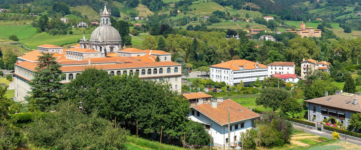A mountainous landscape with a prominent church with a dome and red-tiled roofs, surrounded by lush greenery and scattered buildings.