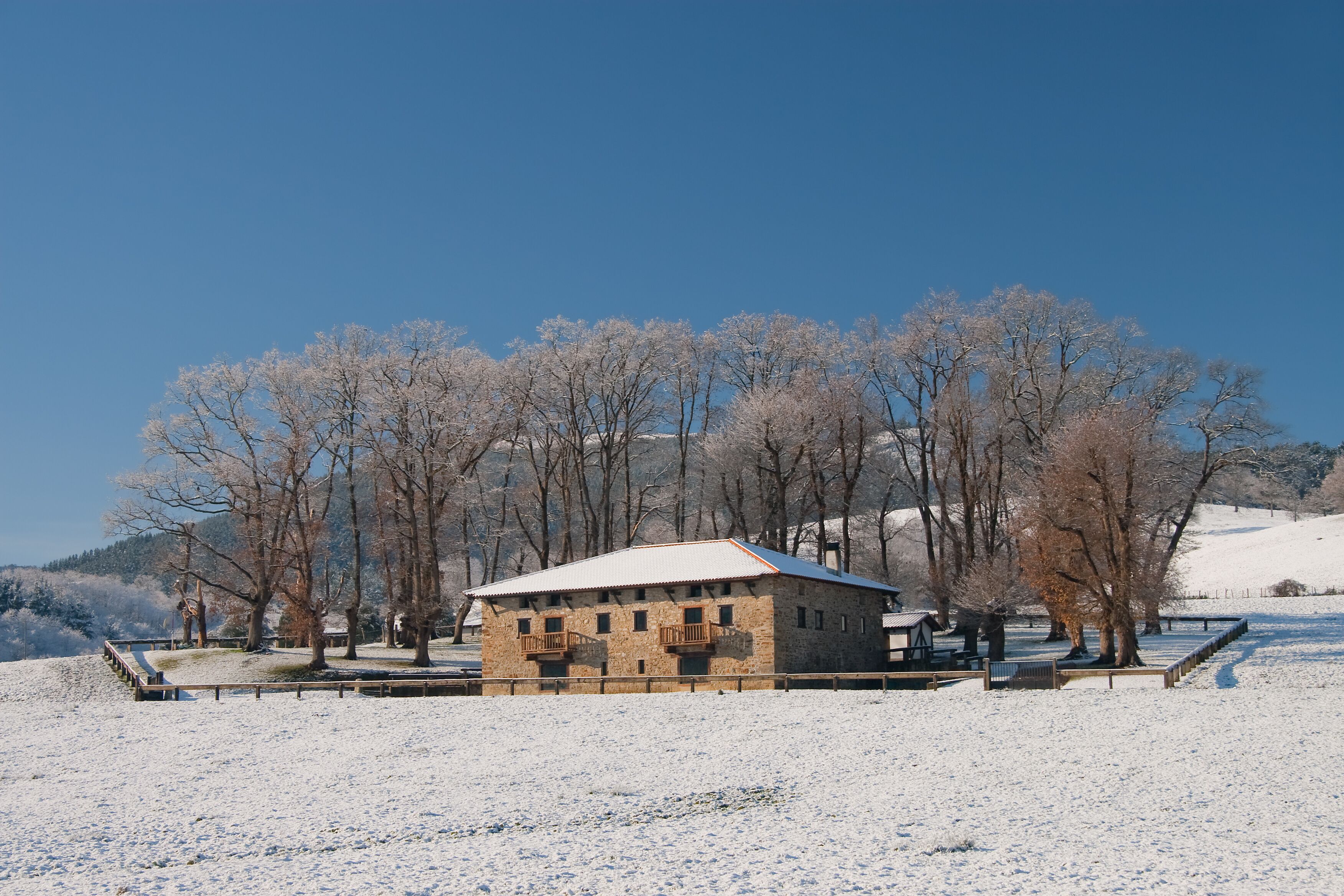 A snow-covered landscape with a stone building, bare trees, and a clear blue sky.