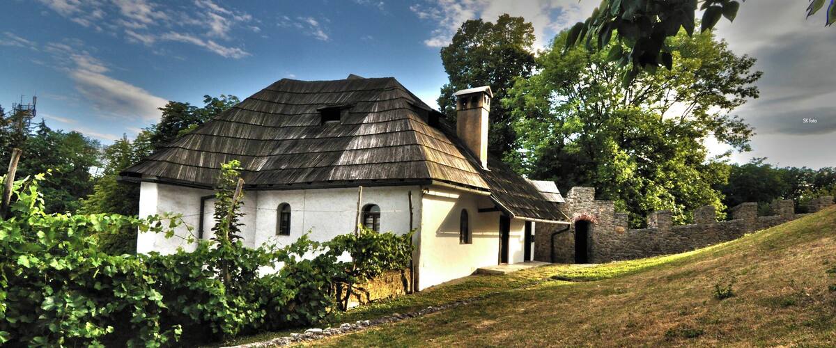 A white house with a dark roof, surrounded by greenery and a stone wall.