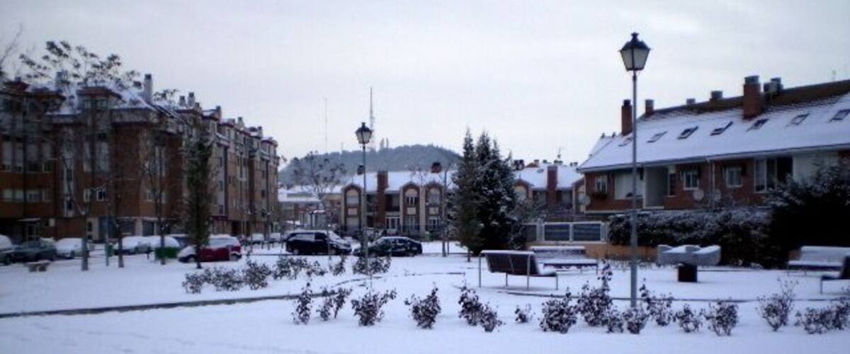 A snowy urban street with parked cars, residential buildings, and a lamppost.