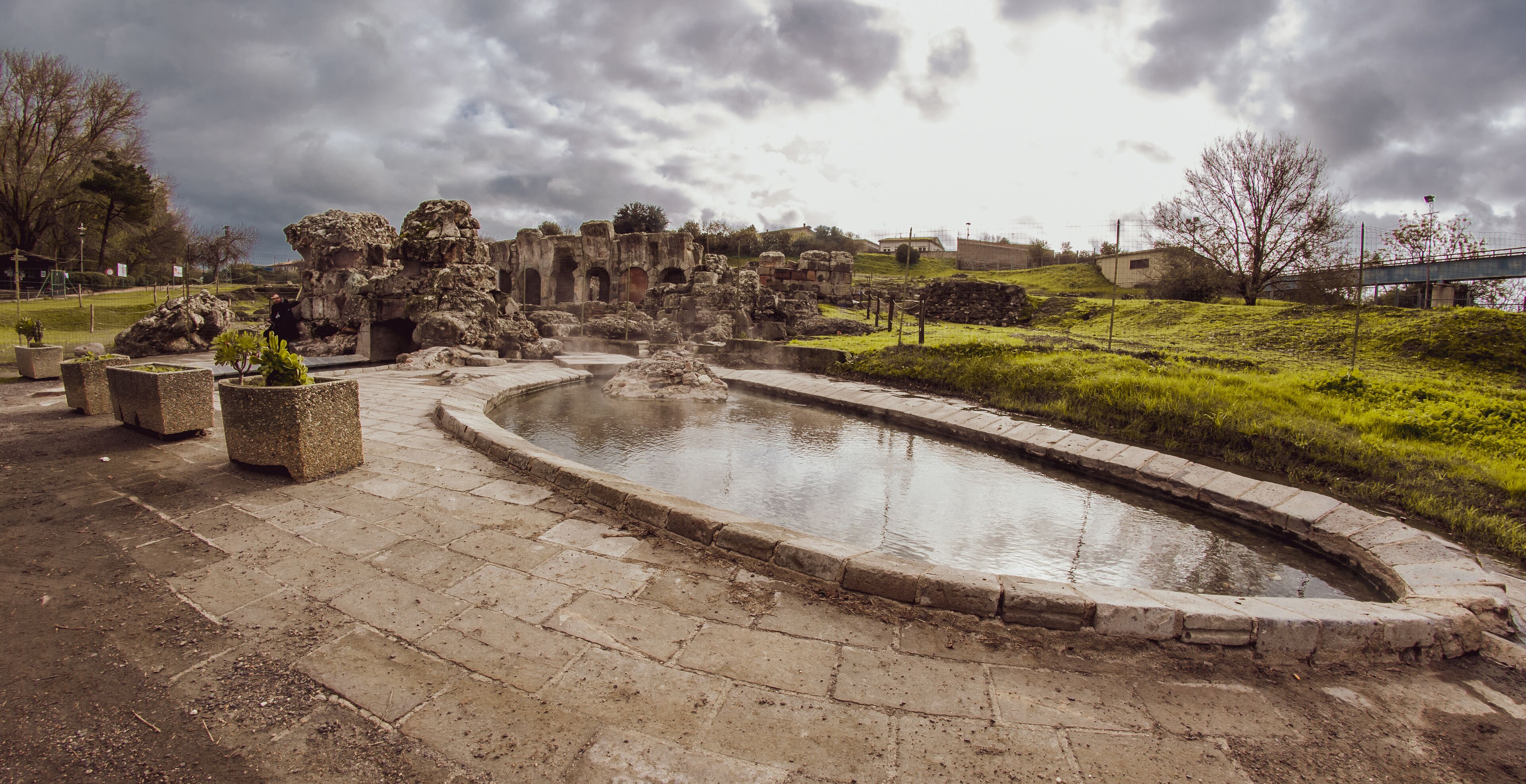Ancient ruins with a central water feature, surrounded by stone pathways and greenery.