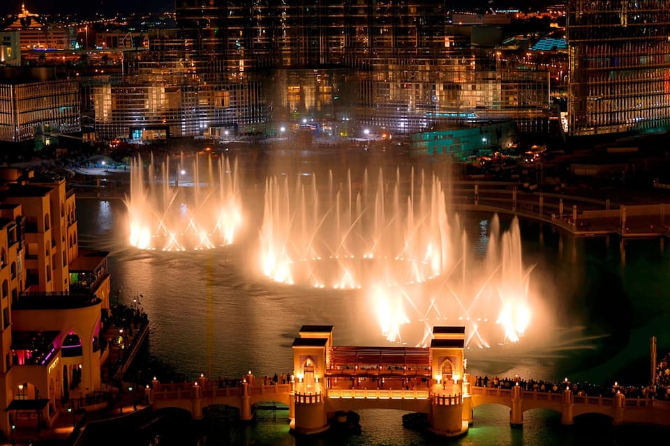 A nighttime cityscape with a large fountain display in the center, surrounded by illuminated buildings and a bridge with people on it.