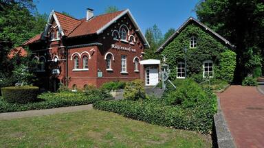 A historic building with a red brick facade and arched windows, surrounded by lush greenery and a clear blue sky.